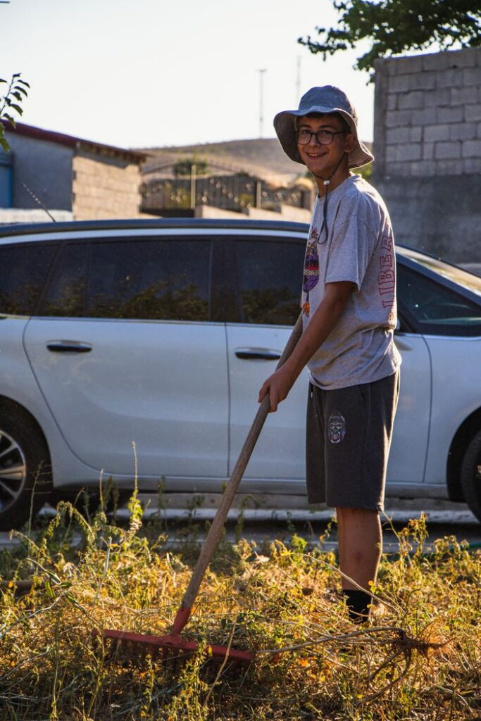 Teen boy wearing bucket hat raking grass in a sunny yard beside a car.